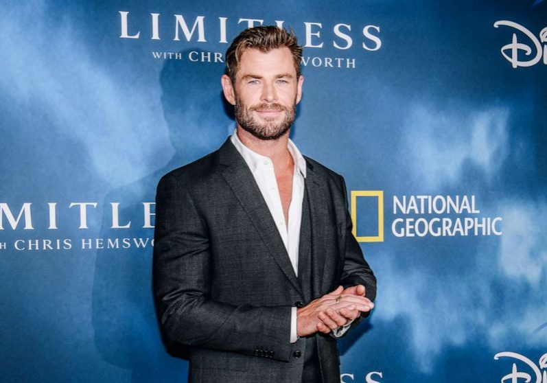 Actor, Chris Hemsworth,  stands in front of a blue sign that advertises the show Limitless.  